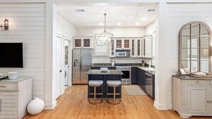 A modern kitchen with white cabinetry, a central island with stools, stainless steel appliances, and a large window with blinds. - WaterColor Park District Retreat, 4BR, Bikes & Walk to Beach (Santa Rosa Beach)