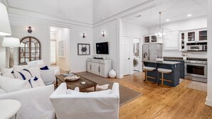 A spacious living room with a large sofa, a coffee table, and a TV. The kitchen area includes a dining table, bar stools, and modern appliances. - WaterColor Park District Retreat, 4BR, Bikes & Walk to Beach (Santa Rosa Beach)