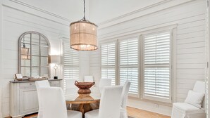 A dining area with a round wooden table, white upholstered chairs, a hanging light fixture, and a large mirror above the table. - WaterColor Park District Retreat, 4BR, Bikes & Walk to Beach (Santa Rosa Beach)
