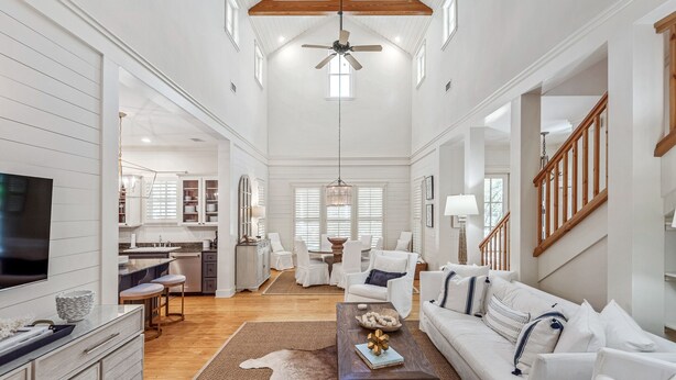 A spacious living room with a high ceiling, wooden beams, a large TV, white furniture, and a dining area with a chandelier. - WaterColor Park District Retreat, 4BR, Bikes & Walk to Beach (Santa Rosa Beach)