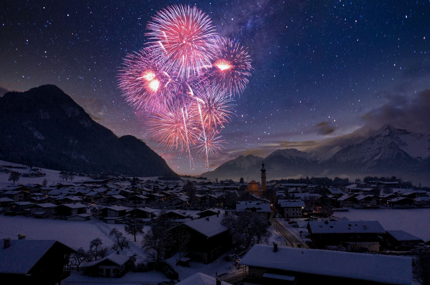 Ein verschneites Dorf mit Häusern und einer Kirche, unter einem Nachthimmel voller Feuerwerk.