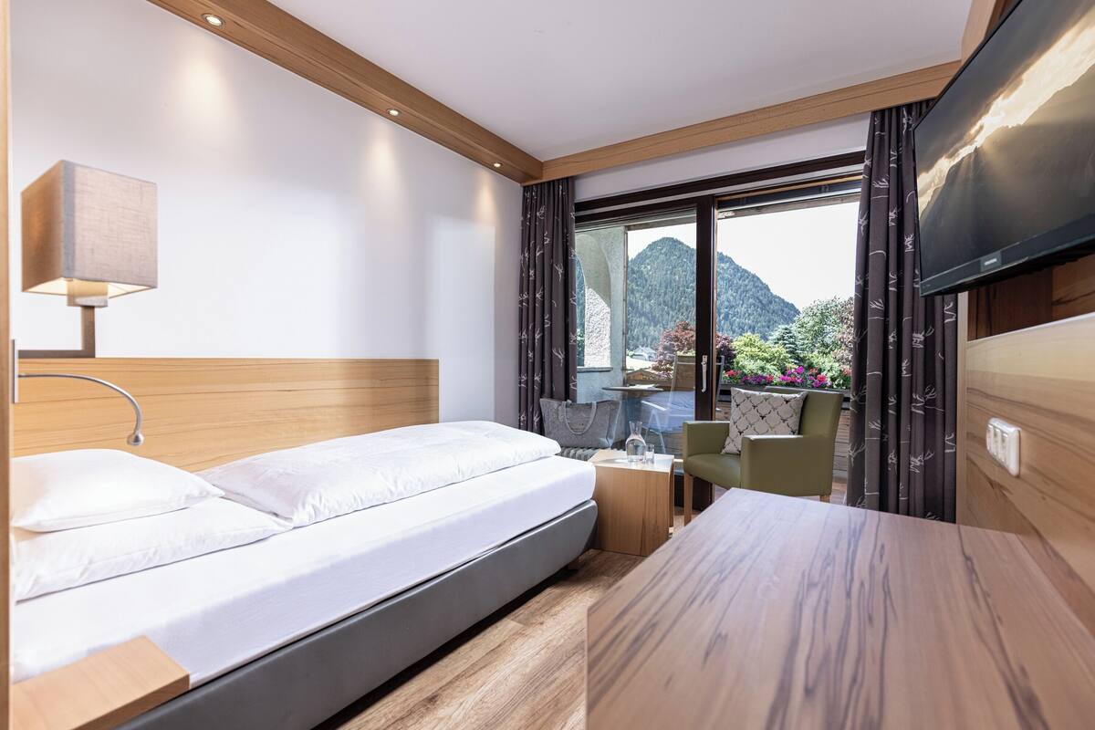 A modern hotel room with a bed, bedside table, lamp, TV, and a view of mountains.