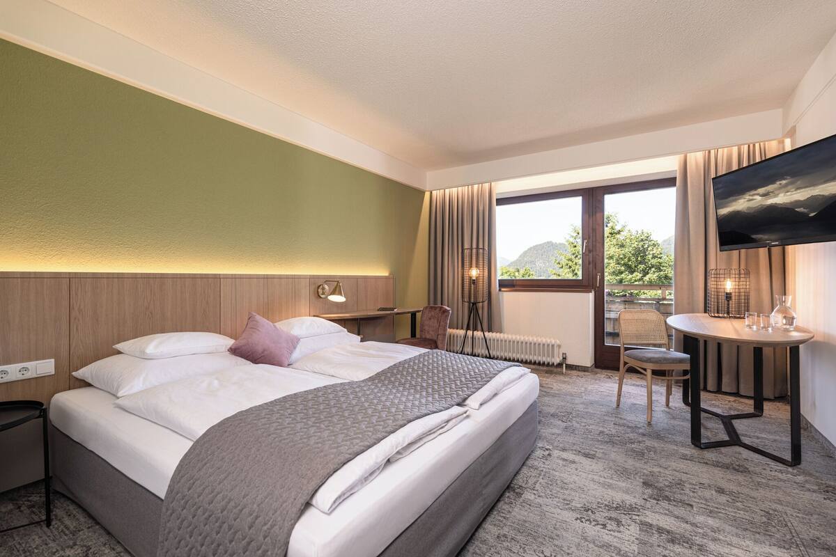 A hotel room with a large bed, a desk, a chair, a TV, and a view of the outdoors.
