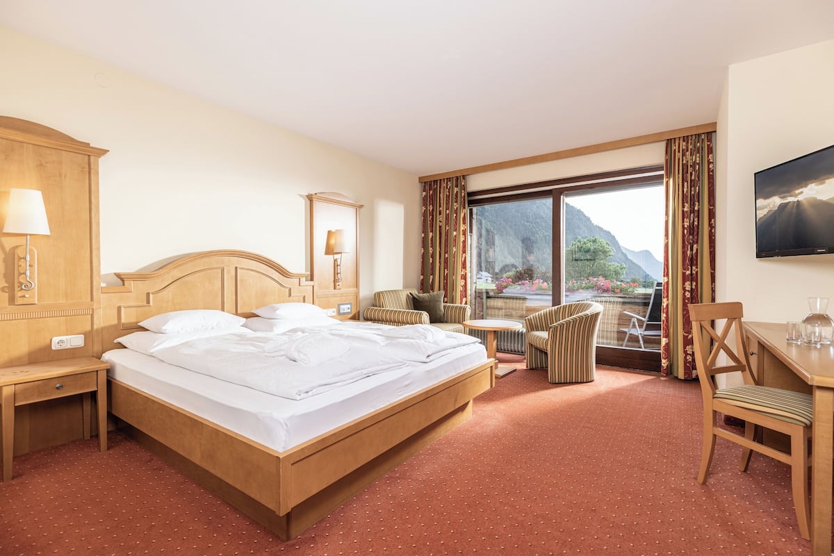 A hotel room with a bed, a desk, a chair, a small table, a lamp, a TV, and a large window with a mountain view.
