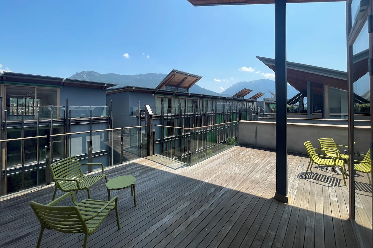 Een houten terras met buitenmeubels, glazen hekwerk en uitzicht op bergen.