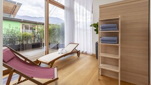A room with a wooden floor, a large window with curtains, a wooden shelf with towels, and a lounge chair. - Habitat Guest House (Trento)