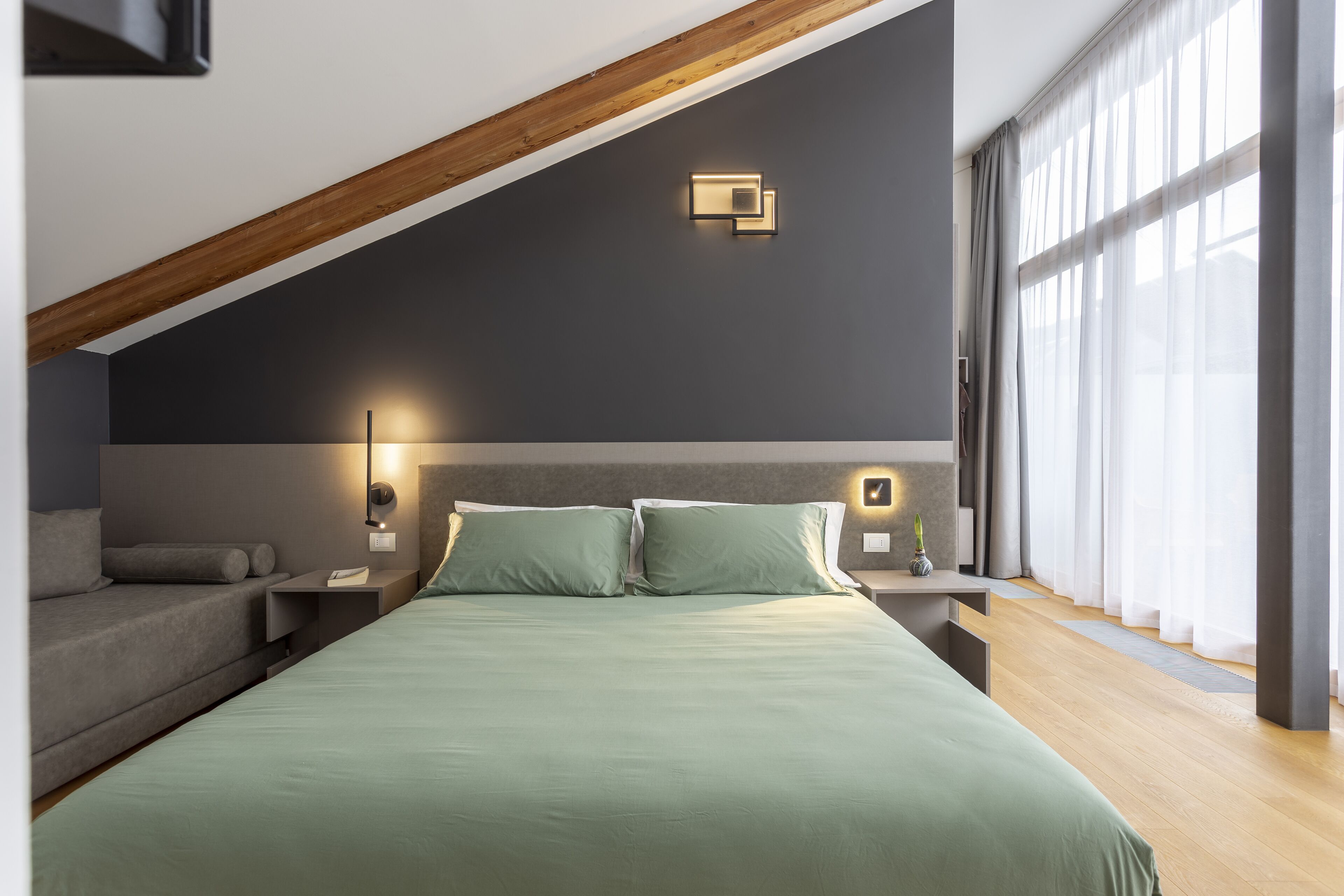 A modern bedroom with a sloped ceiling, a large bed with green bedding, a nightstand with a lamp, and a window with sheer curtains.