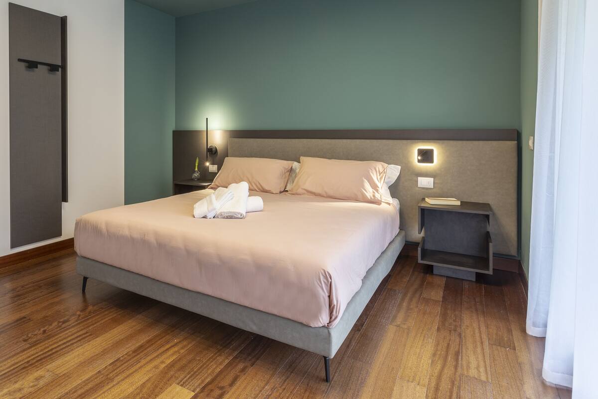 Een net opgemaakt bed met een lichtkleurige dekbedovertrek en kussens, een nachtkastje met een lamp en een klein wandrek.