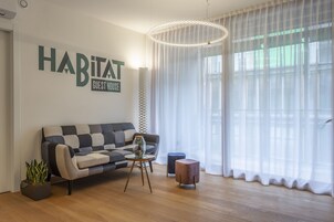 A modern living room with a checkered sofa, a round table, and a potted plant. A sign on the wall reads "HABITAT GUEST HOUSE."
