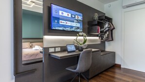 A modern bedroom with a built-in desk, a flat-screen TV, and a bed with a view of the city. - Habitat Guest House (Trento)