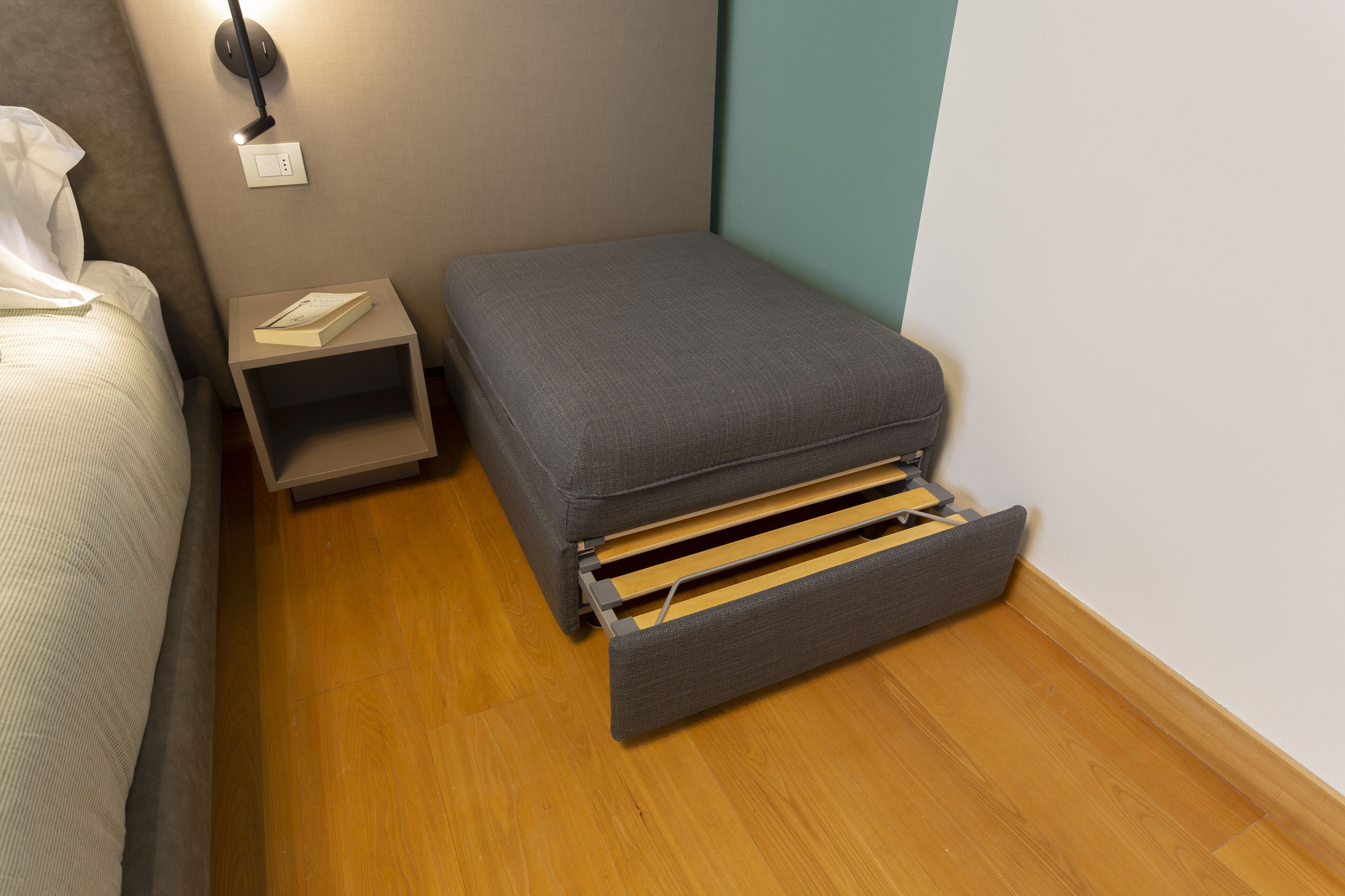 A compact room with a bed, a small bedside table, a nightstand, and a pull-out drawer bench.