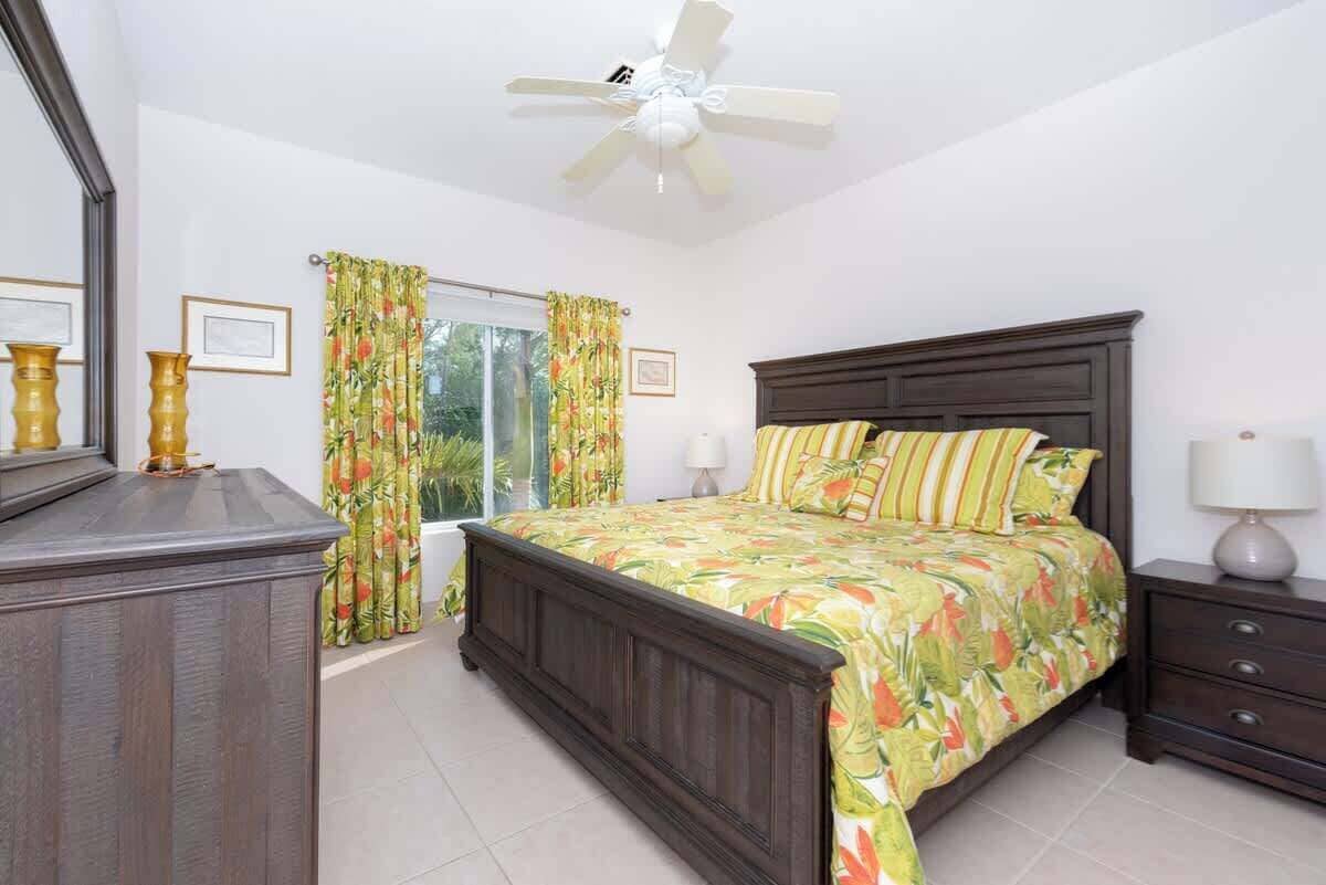 Une chambre à coucher comprenant un lit, une table de chevet, une commode, un ventilateur de plafond et des rideaux.