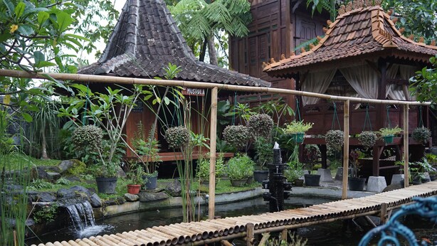 A traditional wooden structure with a tiled roof, surrounded by lush greenery and a small water feature.