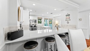 A modern kitchen with white cabinetry, a large island with stools, and a view of the backyard through the windows. - Gulf and Pool Views with Beach Access (Santa Rosa Beach)