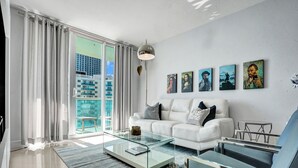 Una sala de estar moderna con un sofá blanco, una mesa de centro de vidrio y vistas a un paisaje urbano a través de amplios ventanales.