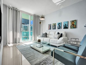 A modern living room with a white sofa, glass coffee table, and a view of a cityscape through large windows.