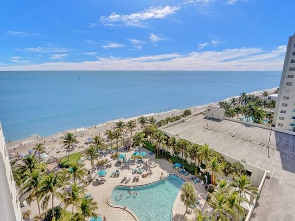 A resort with a swimming pool surrounded by palm trees and sun loungers, overlooking a beach and the ocean.