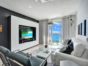 A modern living room with a large flat-screen TV mounted on a white cabinet, a glass coffee table, a white sofa, and a view of the ocean through large windows.