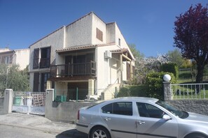 A two-story residential building with a balcony, a car parked in front, and a tree with red leaves.