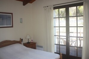 A bedroom with a bed, a nightstand with a lamp, a window with curtains, and a view of an outdoor deck.