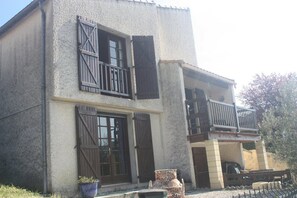 A two-story building with a balcony on the upper floor, a large window on the ground floor, and a potted plant in front.