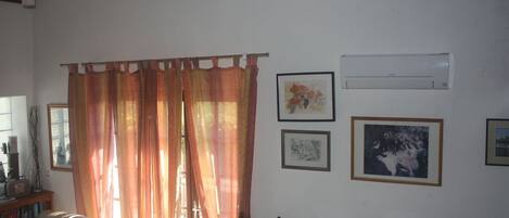Una sala de estar con un sofá, una silla de madera, una estantería y una ventana con cortinas. Hay cuadros enmarcados en la pared.