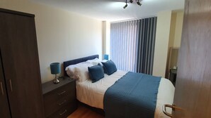 A bedroom with a bed, a wardrobe, a bedside table with a lamp, and a window with blinds. - Manchester City Centre Apartment 1 Bed +sofa Bed (Manchester)