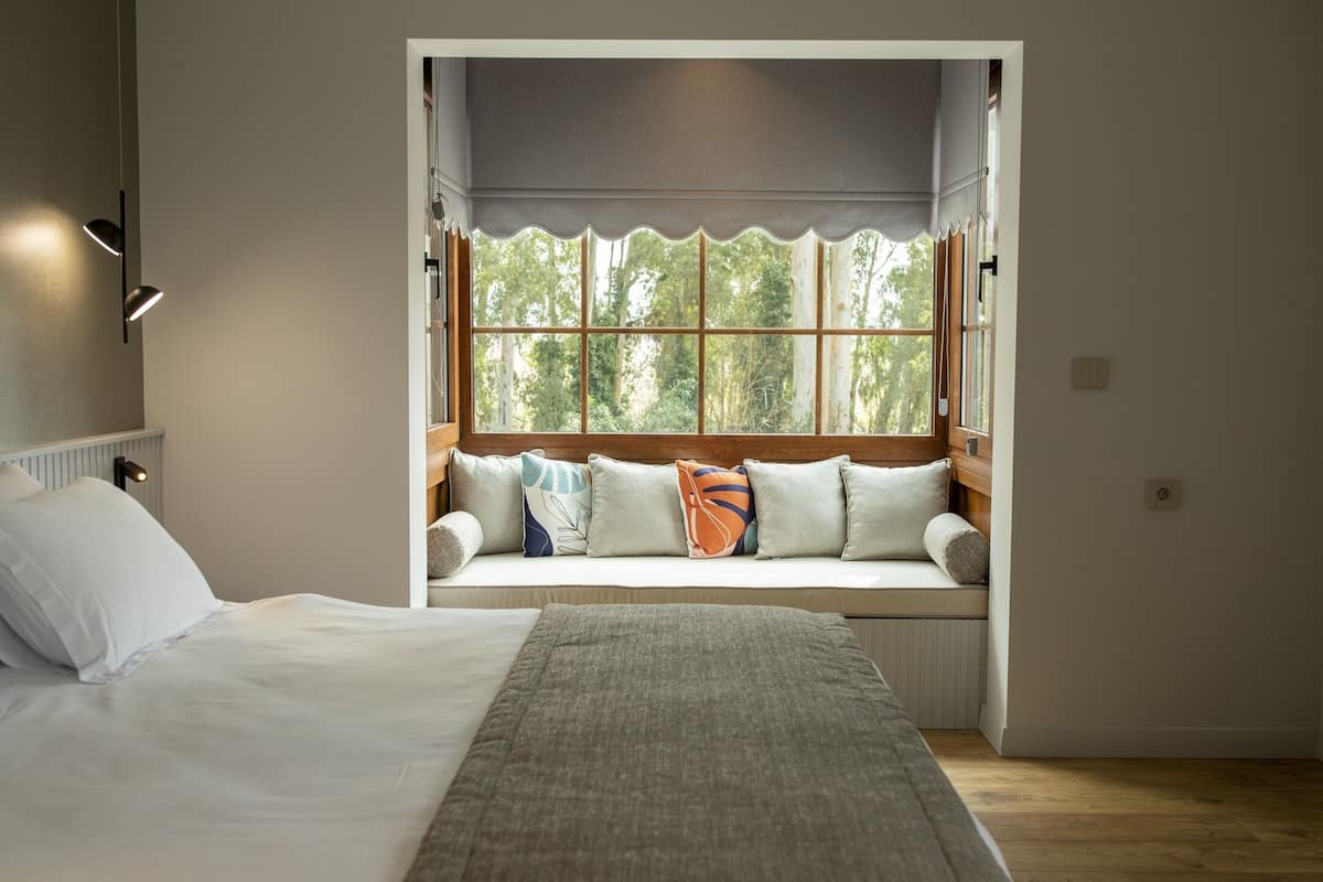 A bedroom with a large window, a bed, a bedside table, and a nightstand.