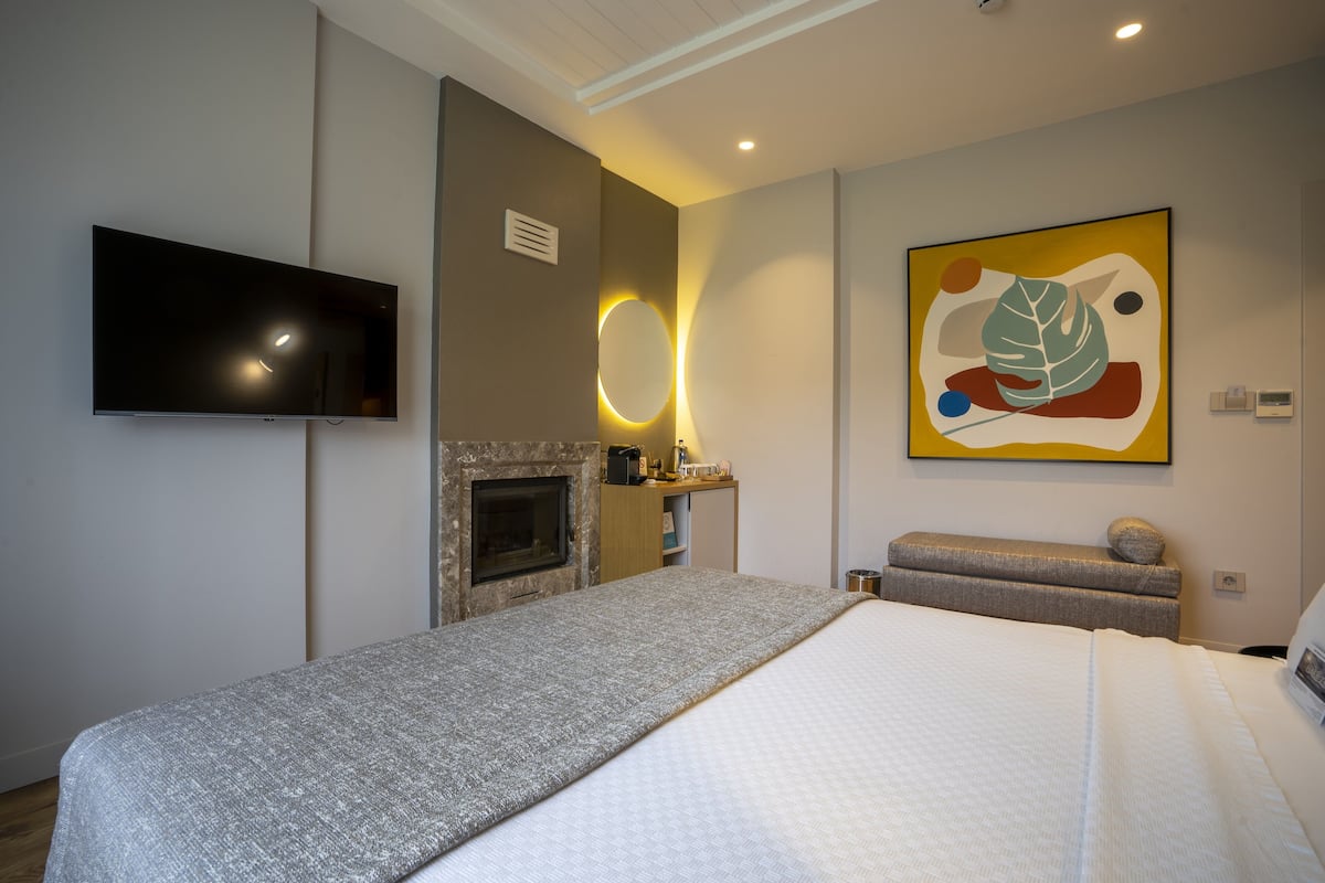 A modern hotel room with a large bed, a flat-screen TV mounted on the wall, a fireplace, a bench, and a colorful abstract painting.