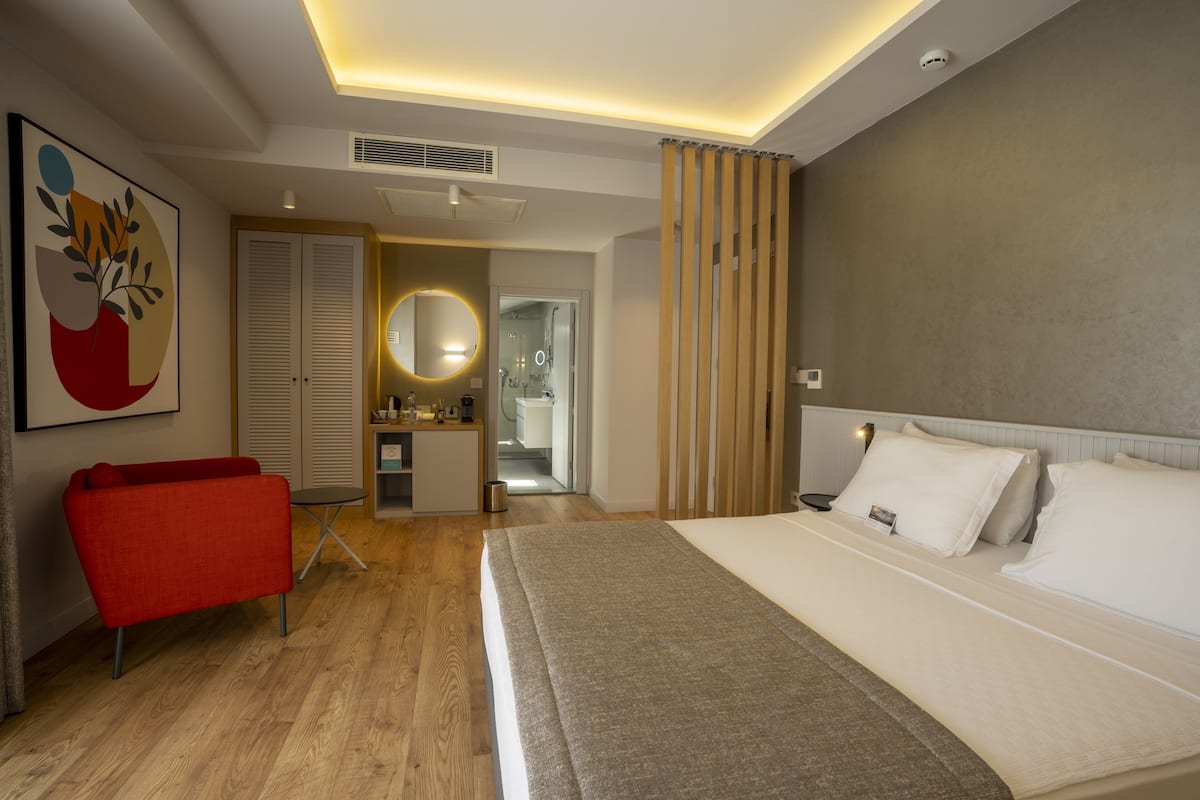 A modern hotel room with a large bed, a red armchair, a bathroom with a mirror, and a ceiling light.