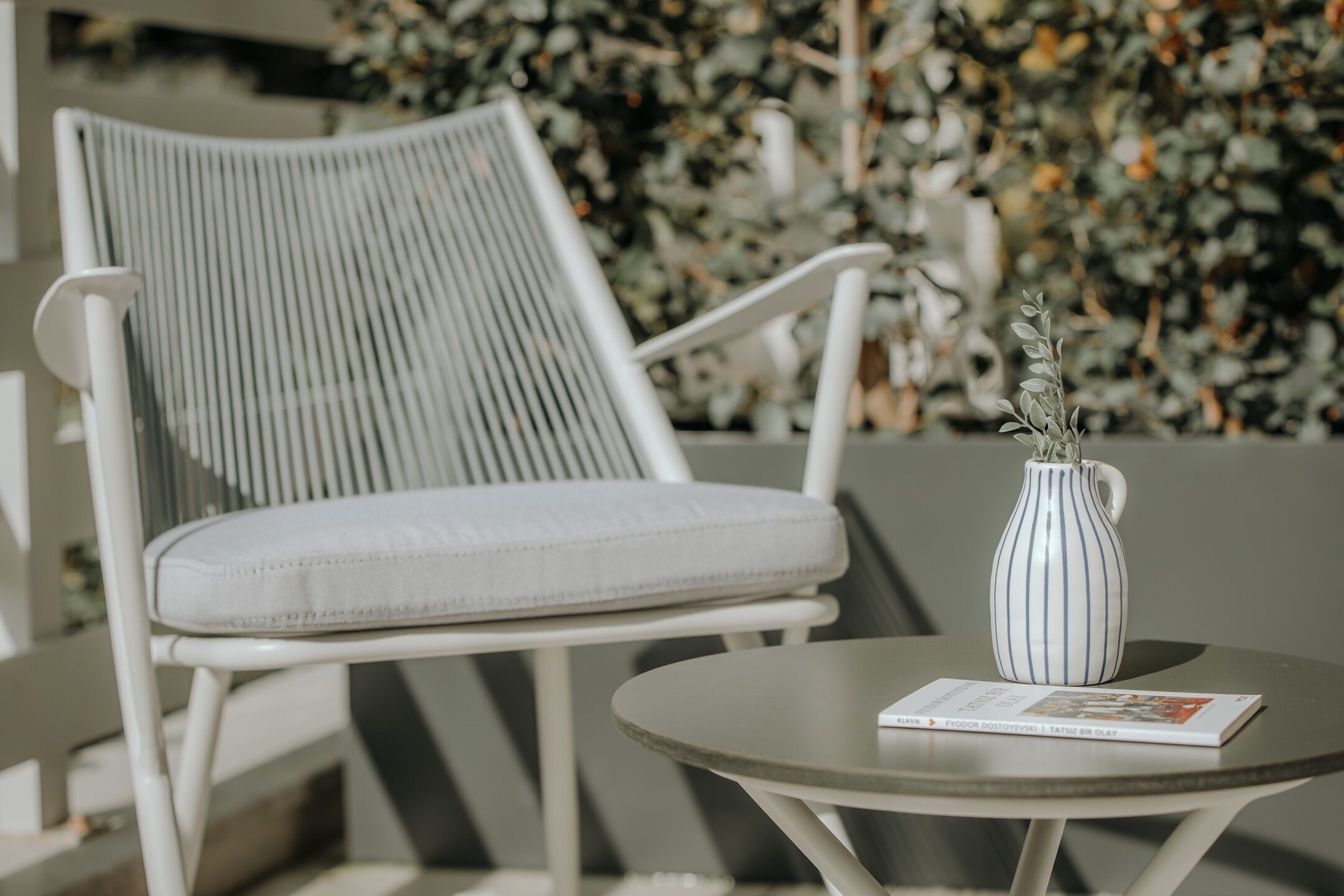 Une chaise blanche avec un dossier à rayures et un siège rembourré, une petite table ronde avec un vase blanc et un magazine, et un décor de feuillage verdoyant.