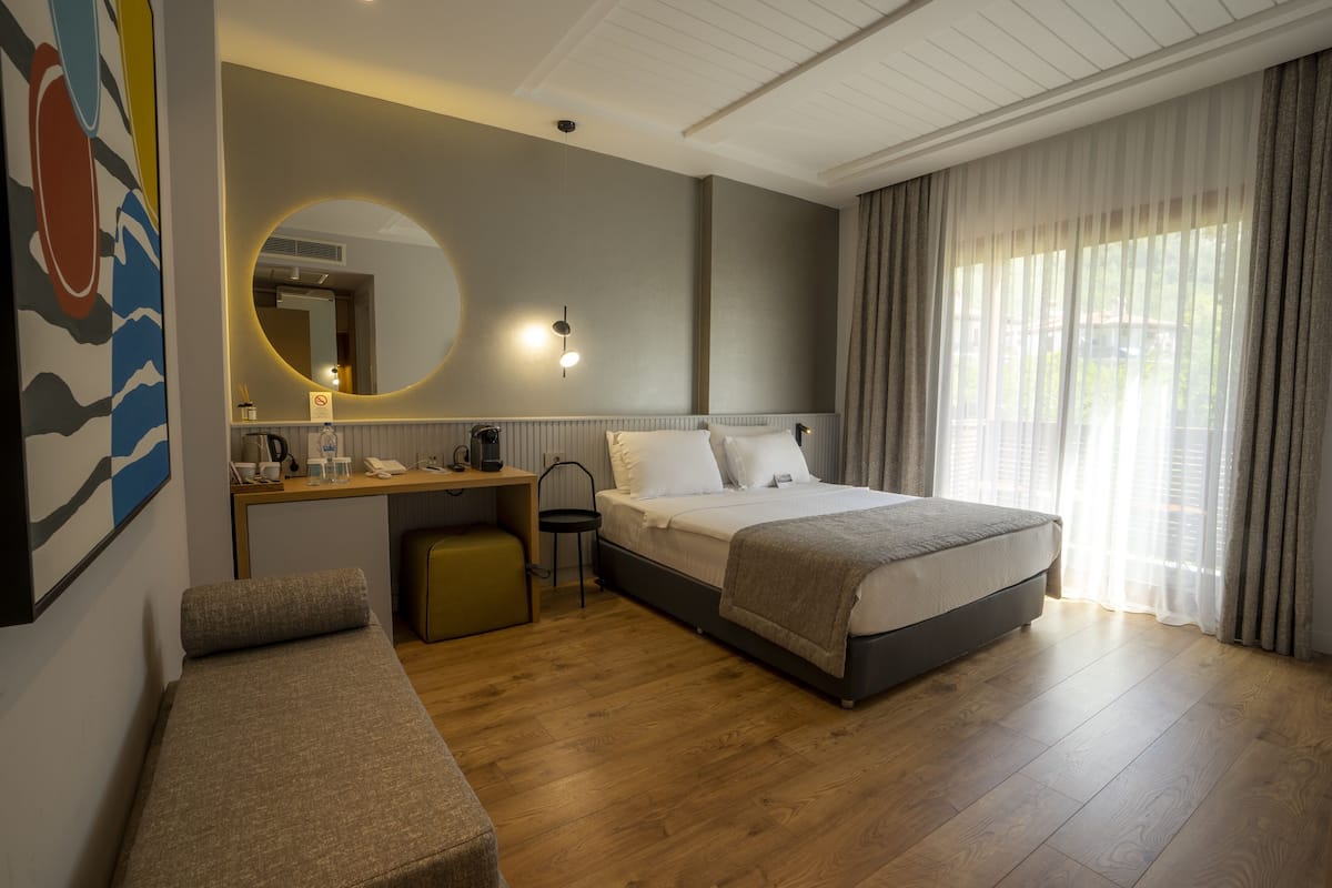 A hotel room with a bed, a desk, a chair, a small bench, a round mirror, and a window with curtains.