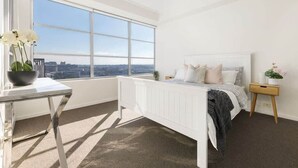 Un dormitorio con un ventanal grande, una cama blanca, una mesita de noche y vistas de la ciudad.