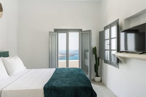 A bedroom with a bed, a TV mounted on the wall, a potted plant, and a view of the sea through a glass door.