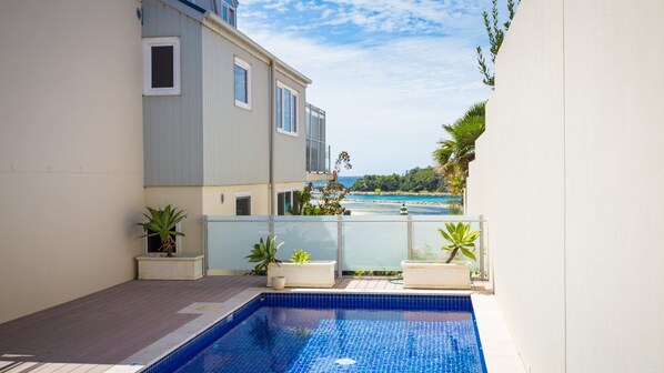 A swimming pool with a view of the sea, surrounded by a building with large windows and potted plants. - Wharf Apartment Unit 11 (NAROOMA)