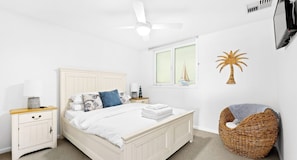 A bedroom with a bed, nightstand, lamp, wall art, and a wicker chair.