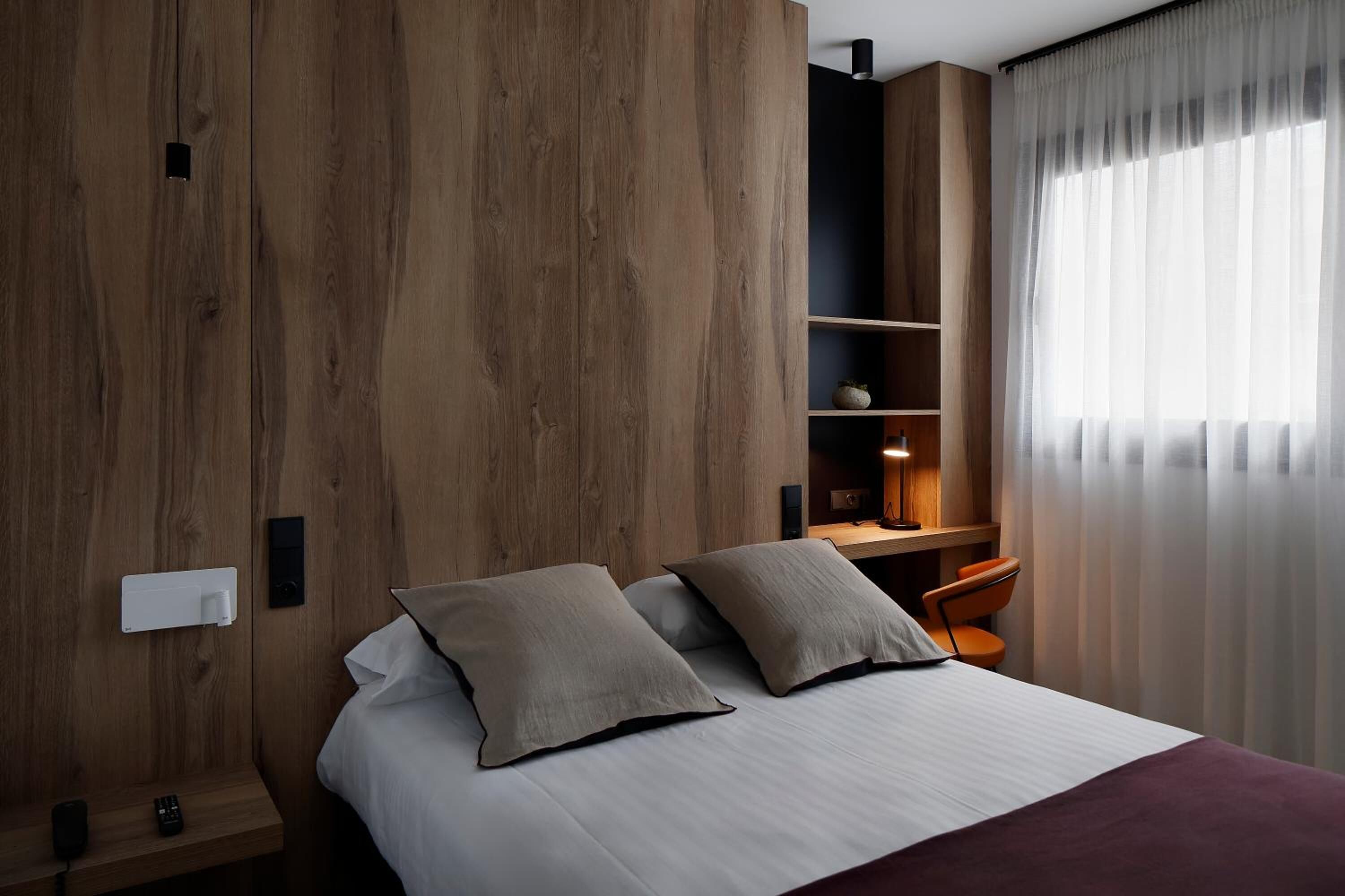 Una habitación de hotel moderna con un muro decorativo de madera, una cama con dos almohadas, un escritorio con una silla y un ventanal con cortinas translúcidas.