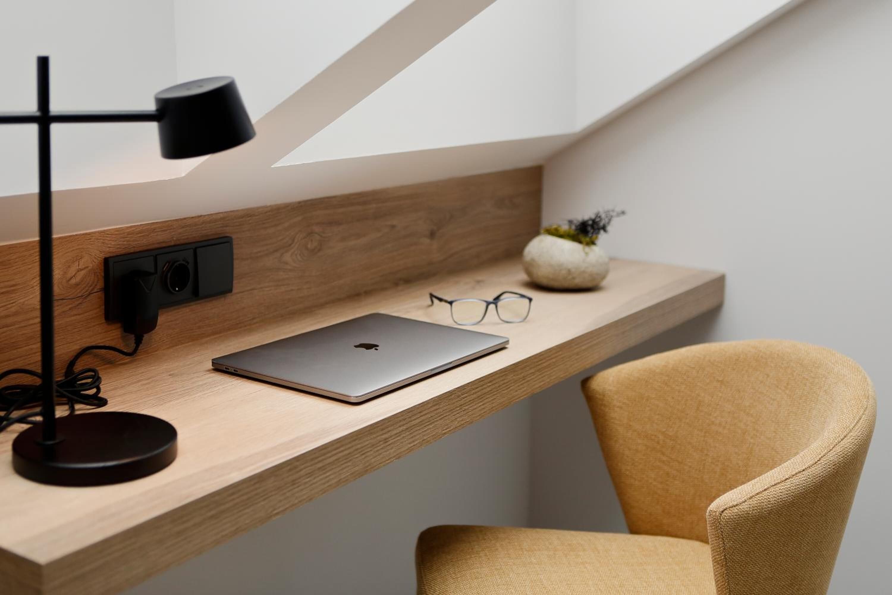 Uma mesa de madeira com um laptop, um par de óculos, uma luminária e um vaso com uma planta.