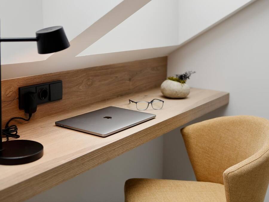 Uma mesa de madeira com um laptop, um par de óculos, uma luminária e um vaso com uma planta.