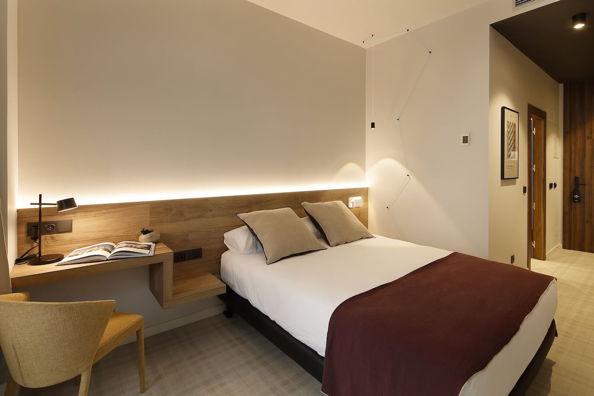 Une chambre d’hôtel moderne, équipée d’un lit, d’une table de chevet, d’une lampe, d’une chaise et d’une étagère murale sur laquelle se trouve un livre.
