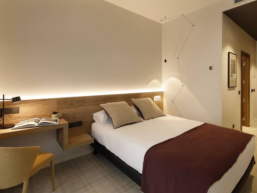 Una habitación de hotel moderna con cama, mesita de noche, lámpara, sillón y estante en la pared con un libro.