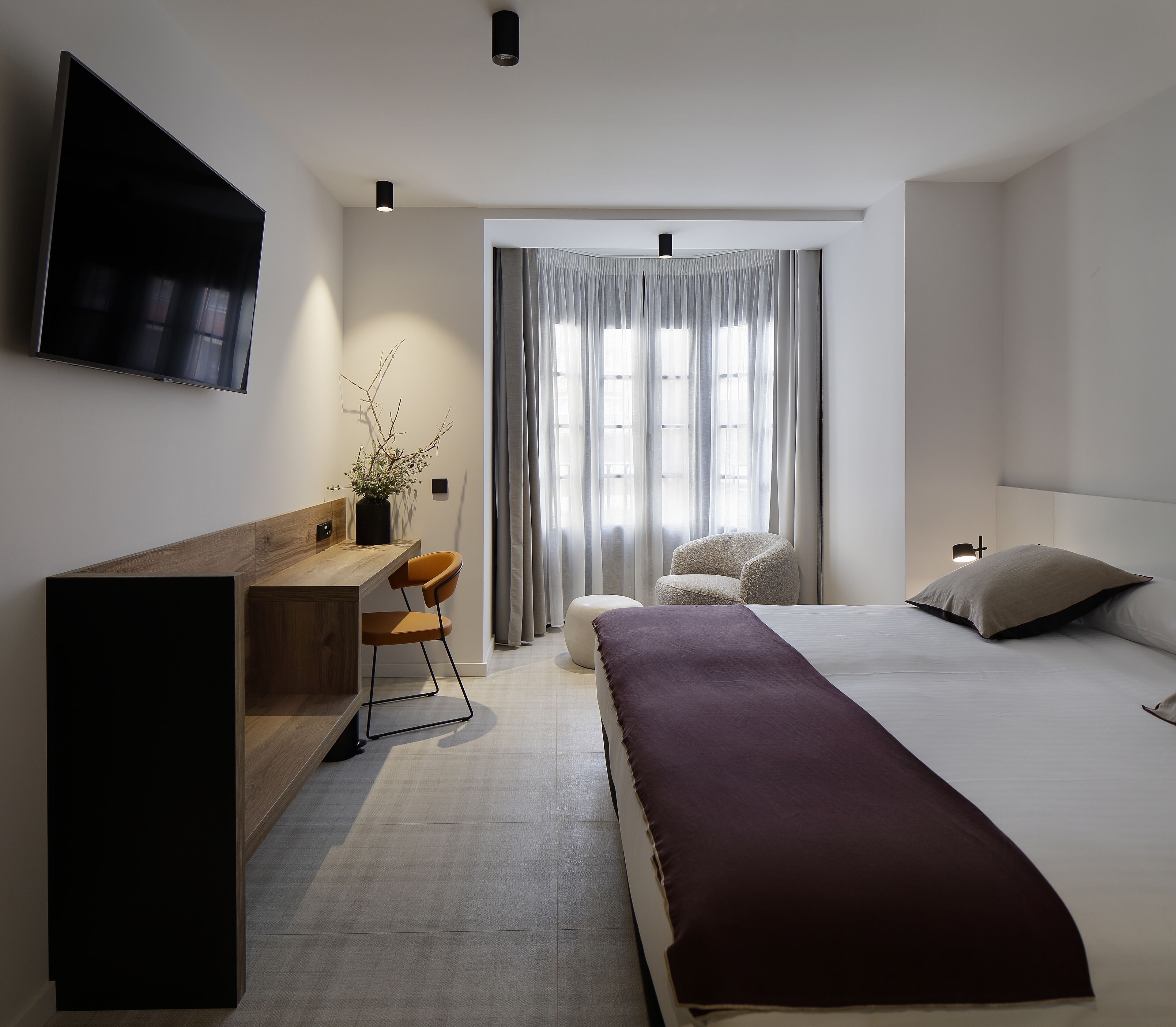 A modern hotel room with a bed, a desk, a chair, a TV, and a window with curtains.