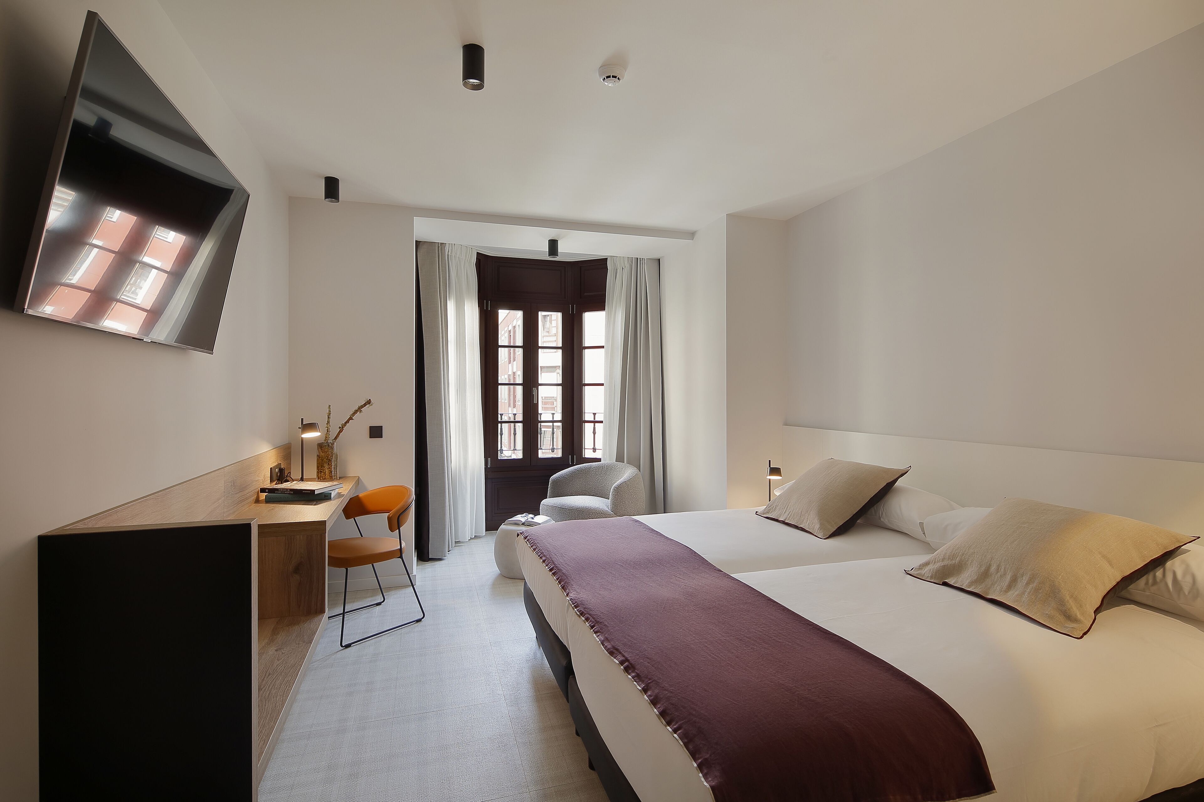 A hotel room with a bed, a desk, a chair, a TV mounted on the wall, and a window with curtains.