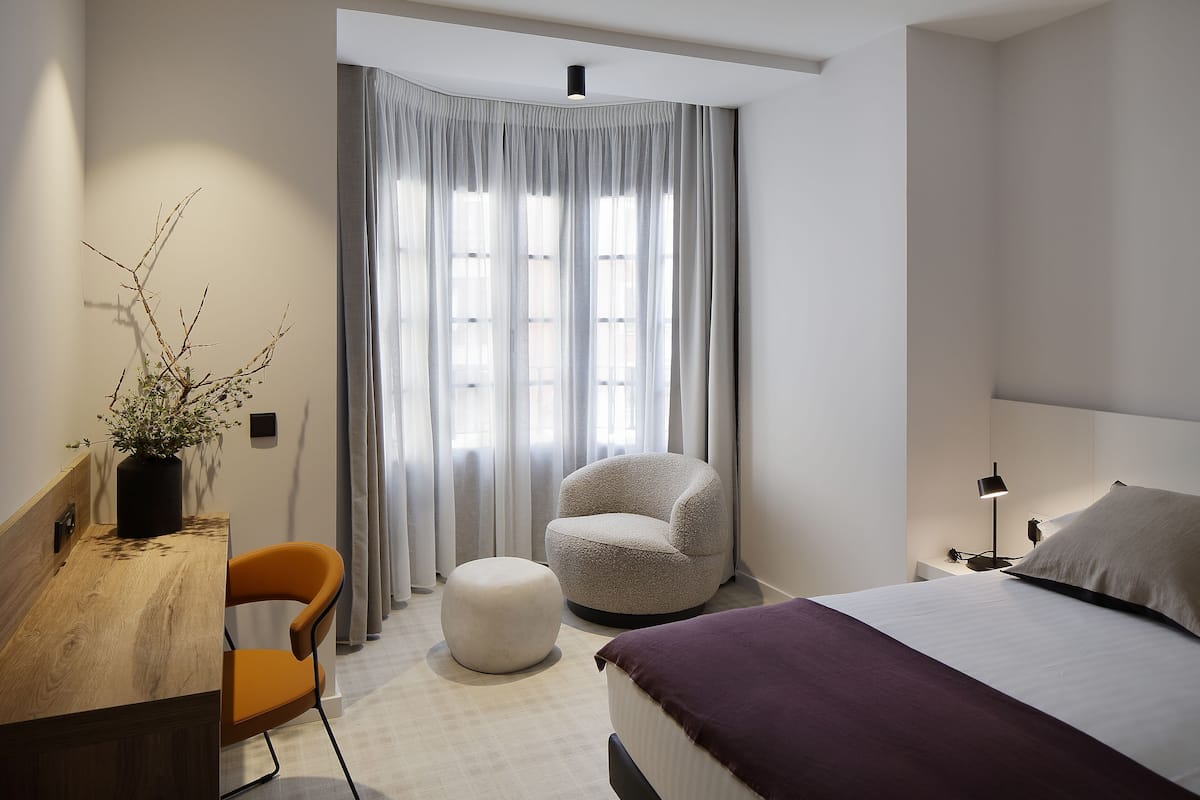 Une chambre moderne avec un bureau en bois, une chaise orange, un pouf rond, un lit recouvert d’un plaid violet et une fenêtre agrémentée de rideaux légers.