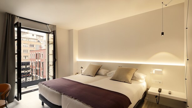 Ein Hotelzimmer mit einem großen Bett, einem Nachttisch, einem Telefon und Blick auf die Stadt durch eine geöffnete Tür.