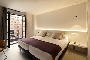 Un dormitorio de hotel con una cama grande, una mesita de noche con lámpara y vista a la ciudad desde una puerta abierta.