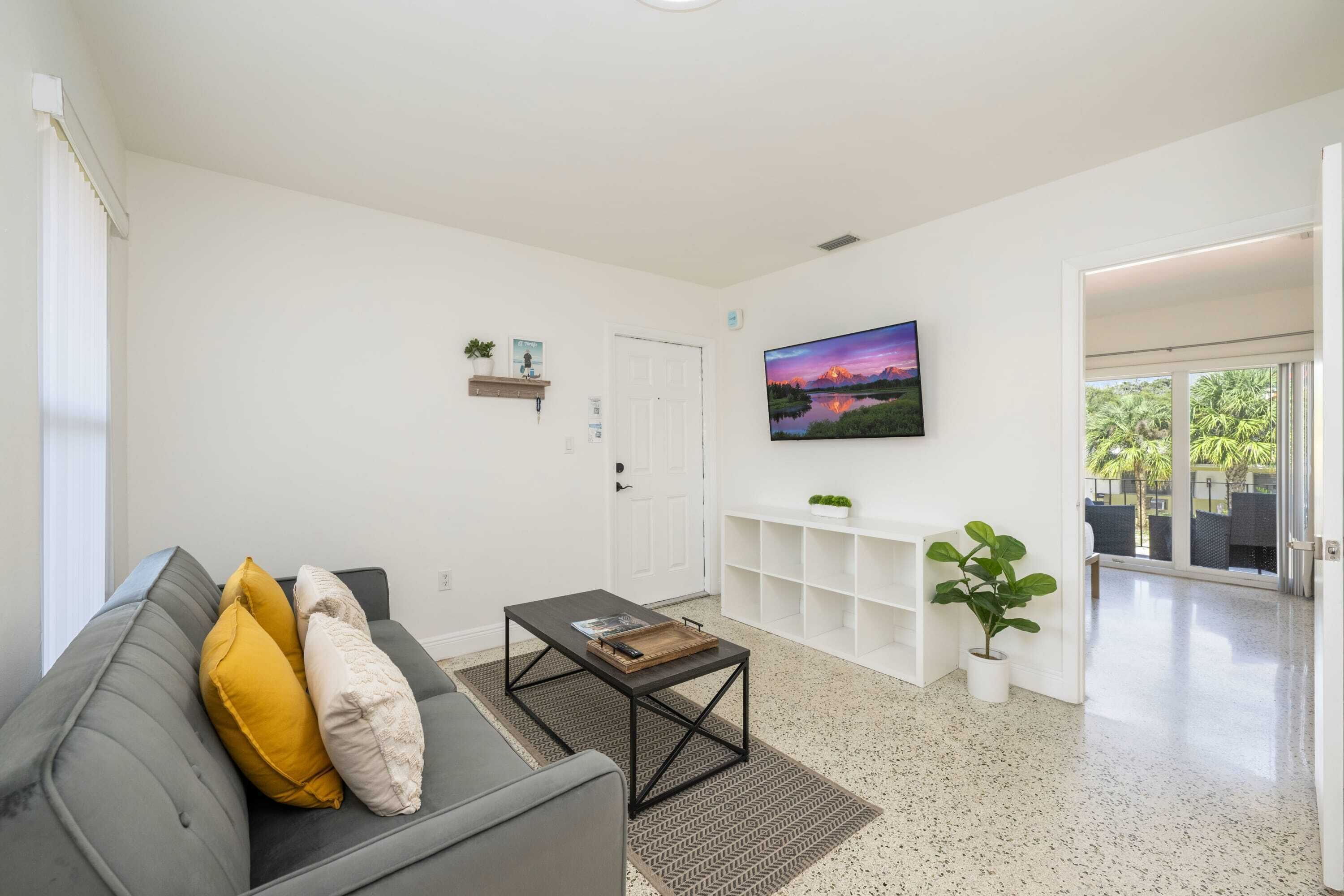 A modern living room with a grey sofa, a coffee table, a TV mounted on the wall, and a door leading to an outdoor area.