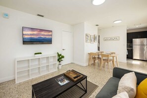 A modern living room with a flat-screen TV, a coffee table, a sofa, and a dining area with a table and chairs.