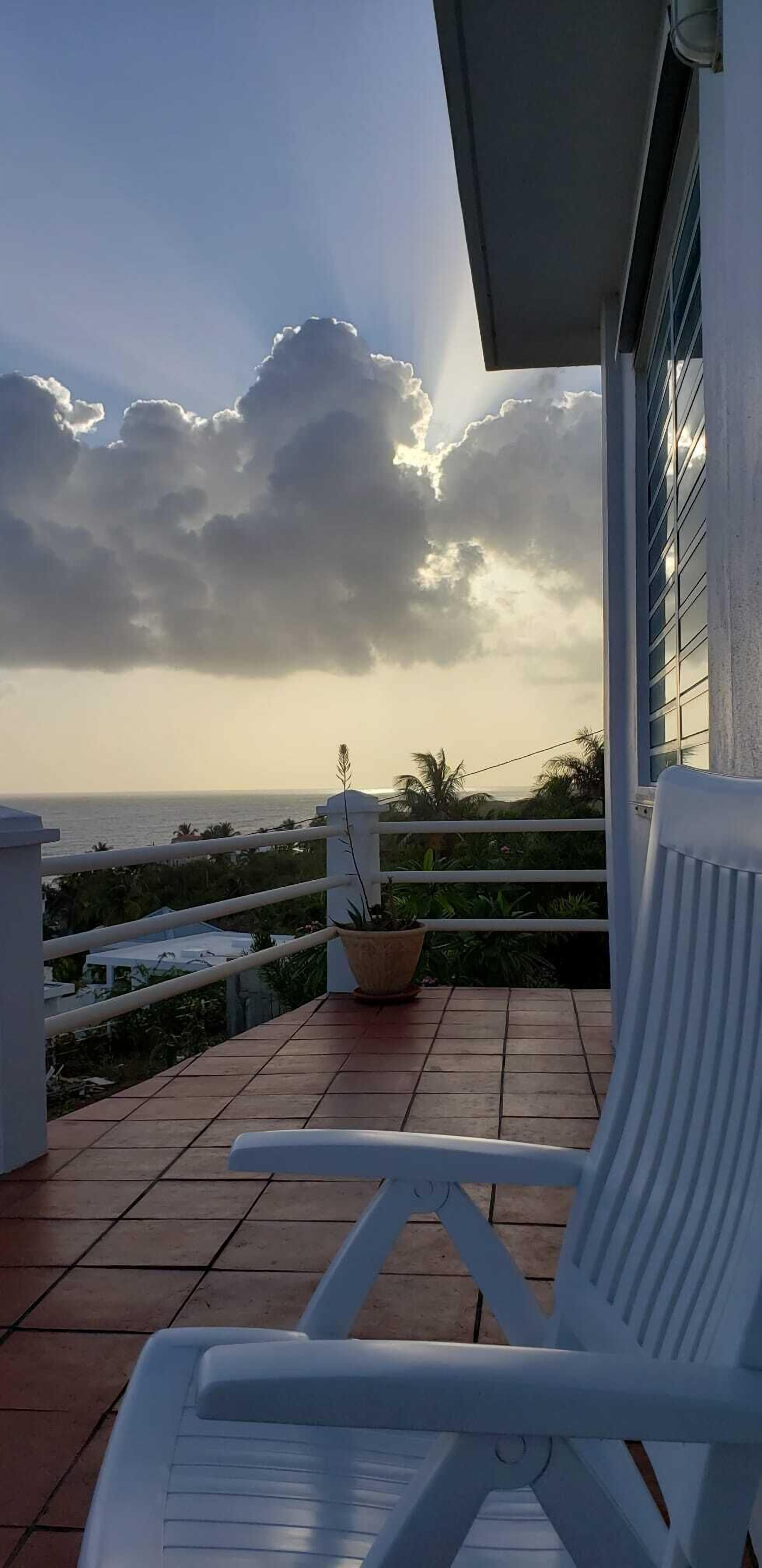 Un balcone con una sedia bianca, una pianta in vaso e vista sull'oceano sotto un cielo nuvoloso.
