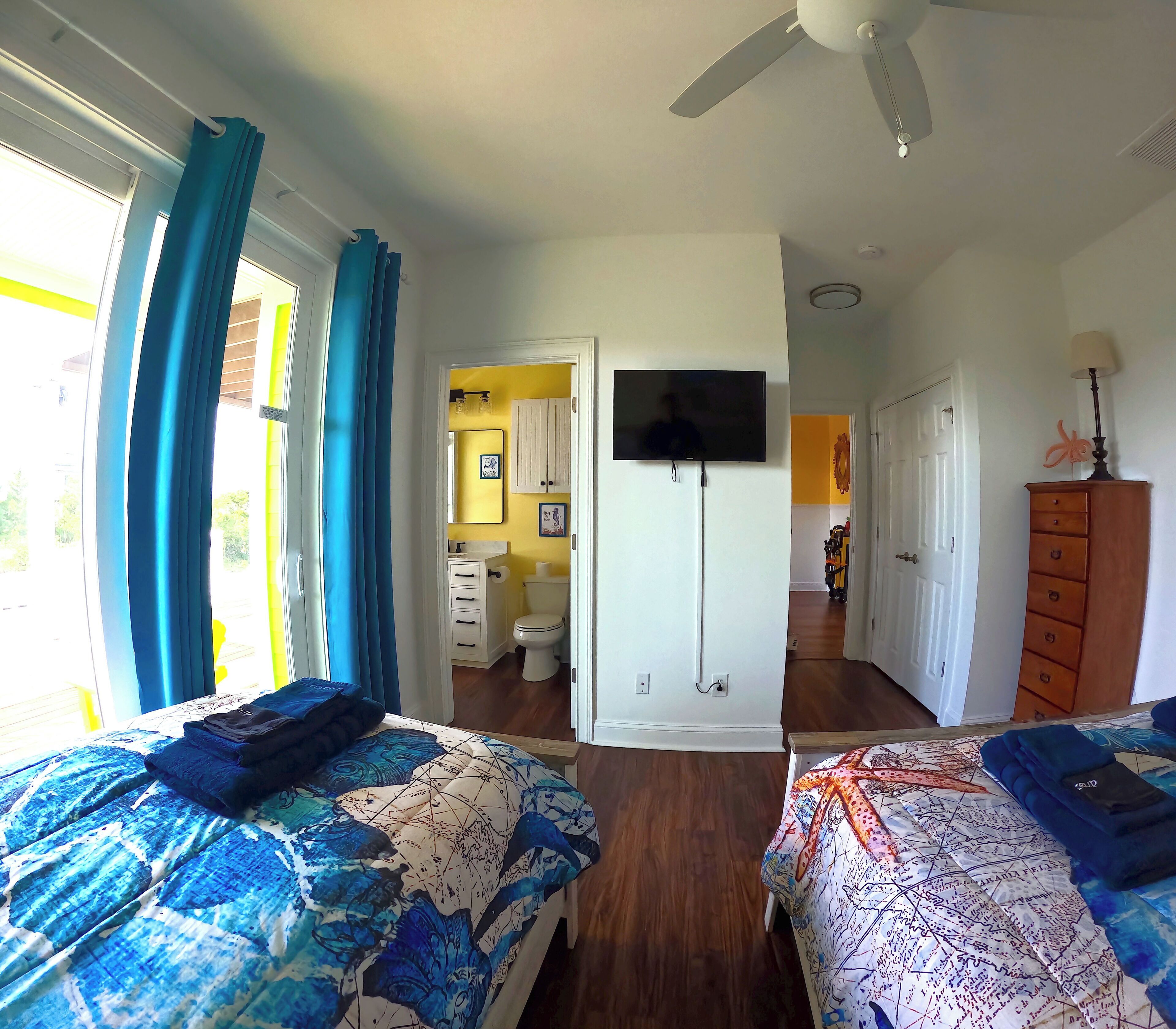 A bedroom with two beds, a TV, a ceiling fan, and a bathroom visible through an open door.